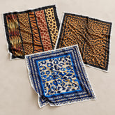 Three square patterned scarves laid flat on a light beige background. One scarf features vertical stripes with tiger, leopard, and giraffe prints in orange, brown, and black.