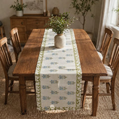 A rectangular wooden dining table in a warm, rustic room setting, styled with a cream table runner featuring a blue and green floral pattern and a matching border. A rustic ceramic vase with green foliage sits in the center of the table, surrounded by wooden chairs.