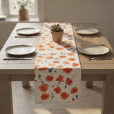 A floral table runner with orange poppy designs and a white base is styled on a light wood dining table, flanked by plates and cutlery in a bright, neutral room.