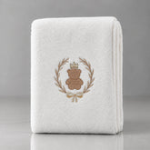 Folded white blanket with a crowned brown teddy bear inside a gold-tone leafy wreath