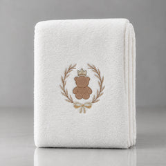 Folded white blanket with a crowned brown teddy bear inside a gold-tone leafy wreath