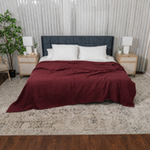 A bed dressed in a textured burgundy blanket and white pillows, positioned in a modern bedroom setting with a dark blue tufted headboard, wooden nightstands, and a patterned area rug.