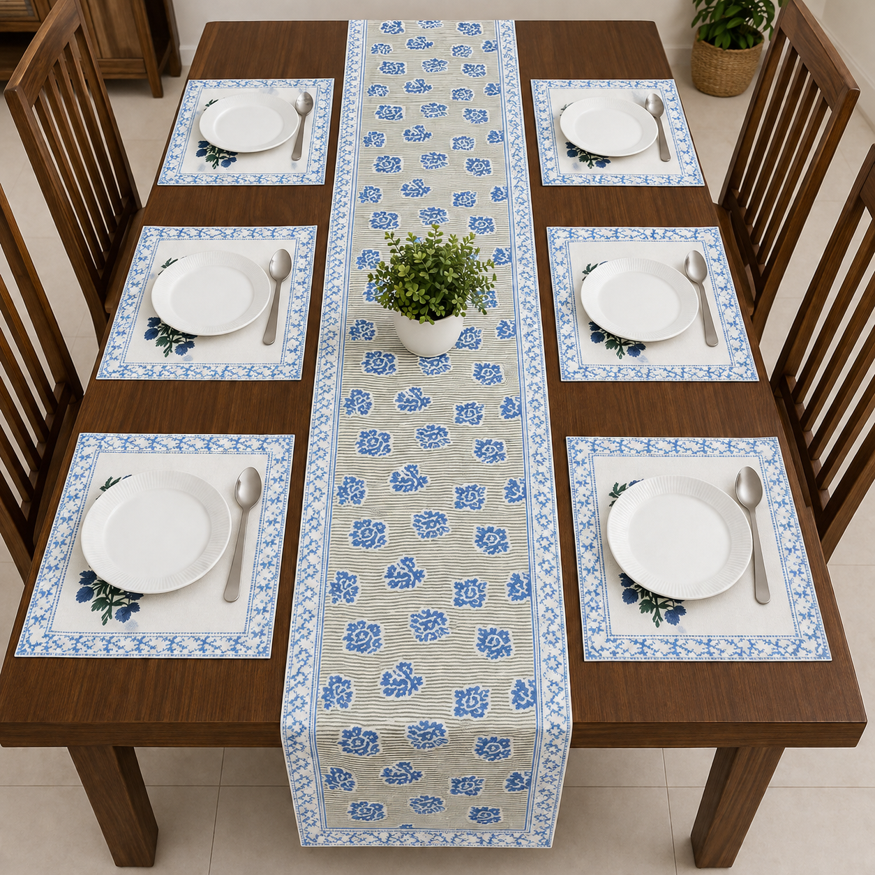 A dining table set with a blue and beige floral table runner down the center, matching placemats with white plates and silverware, and wooden chairs around it.