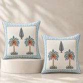 Two decorative square throw pillows with embroidered palm tree motifs and striped blue borders, displayed on a white pedestal against a warm beige background with soft leaf shadows.
