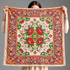 A person holds up a square silk scarf with a vibrant pomegranate and floral pattern, showing the product clearly against a solid gray studio background.