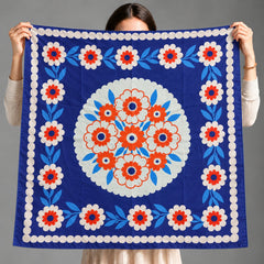 A person holds up a square blue scarf with a white, red, and light blue floral pattern, showing the front design against a plain gray background.