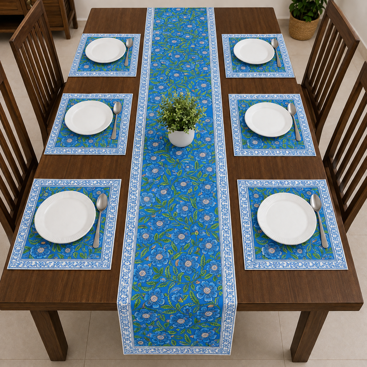 A dining table set with a blue floral table runner down the center and matching blue placemats under six white plates, surrounded by wooden chairs.
