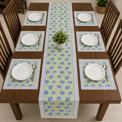 A rectangular wooden dining table set with six matching placemats and a long floral table runner. Each place setting includes a white plate, silver spoon, and folded green napkin, with a small potted green plant centered on the runner.