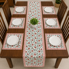 A rectangular wooden dining table set for six with a floral table runner and matching placemats. The placemats and runner feature a red and green floral pattern on a white background with red-and-white striped borders.