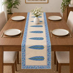 Wooden dining table set with white plates and gold cutlery, featuring a cream-colored table runner adorned with a line of blue and green textured feathers and a blue floral border.