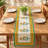 A rectangular table runner with a yellow and green floral pattern is laid down the center of a wooden dining table set with white plates and gold cutlery. A small potted green plant sits on top of the runner.