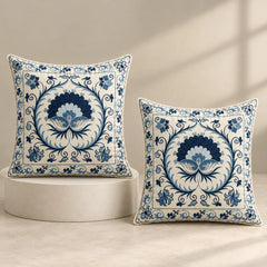 Two square decorative throw pillows with blue and white floral motifs, styled on a neutral beige background with one resting on a round pedestal and the other standing beside it.