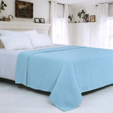 A light blue textured waffle-weave blanket draped over a bed with white sheets and pillows in a bright, white bedroom.