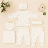 A baby clothing set arranged on a light beige background, including a cream long-sleeve top, matching pants, a hat, a bib, a folded blanket, a folded cloth, and a folded headband, with small animal prints. Decorative props include a plush bunny, a leaf, and a folded leaf in the corners.