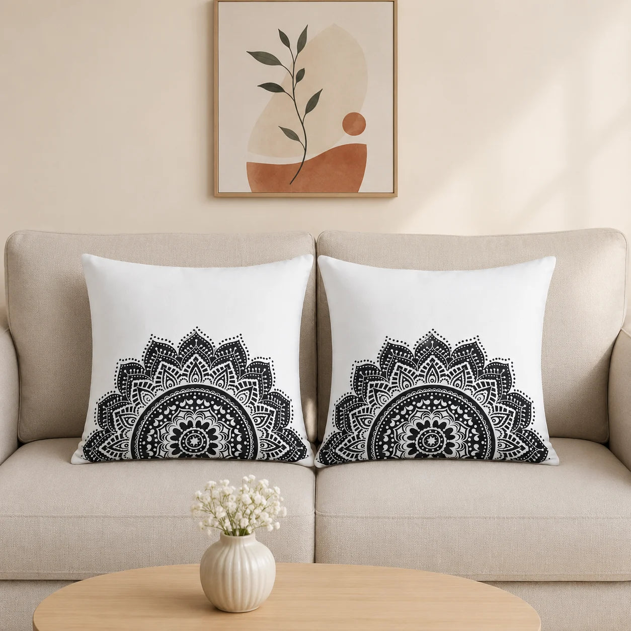 Two square white throw pillows with black mandala patterns, styled on a beige sofa in a living room setting with a small table and vase in the foreground and framed wall art above.