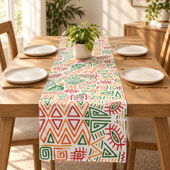 A colorful table runner with a geometric tribal pattern in green, orange, red, and yellow on a white background, styled on a wooden dining table with plates and a small potted plant.