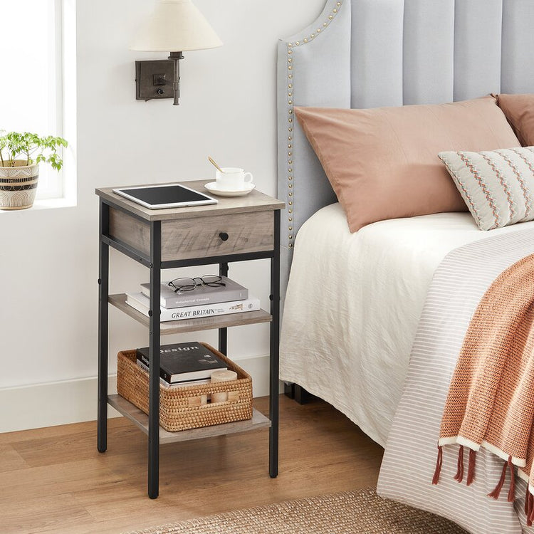 Narrow gray-brown side table with drawer and shelves beside a bed