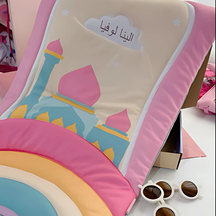 Pastel children's prayer mat with a mosque and rainbow design.
