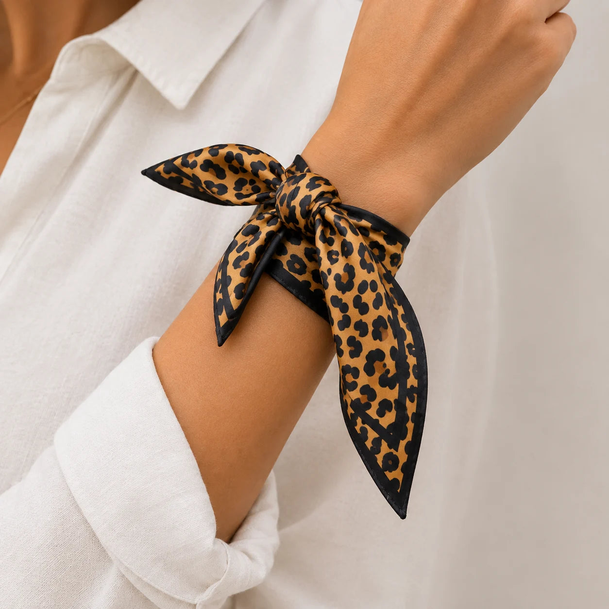 A leopard print silk scarf tied in a knot around a woman's wrist, worn with a white collared shirt.
