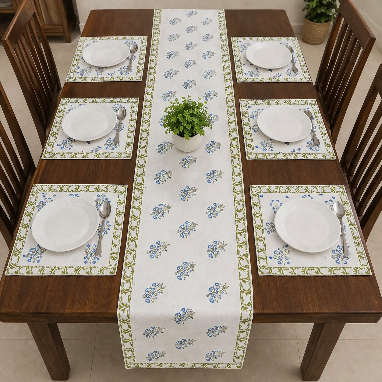 A rectangular dining table set with six white plates, silver spoons and forks, matching placemats, and a long white table runner printed with a blue floral pattern and green border. The table is surrounded by wooden chairs, with a small potted green plant in the center.