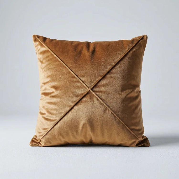 A square velvet throw pillow in a warm camel brown color, featuring a central crisscross design that forms an X shape. The pillow is photographed against a neutral light gray background.