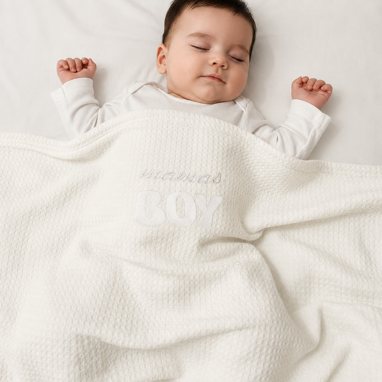 Infant under a white waffle-textured blanket with white mamas BOY lettering