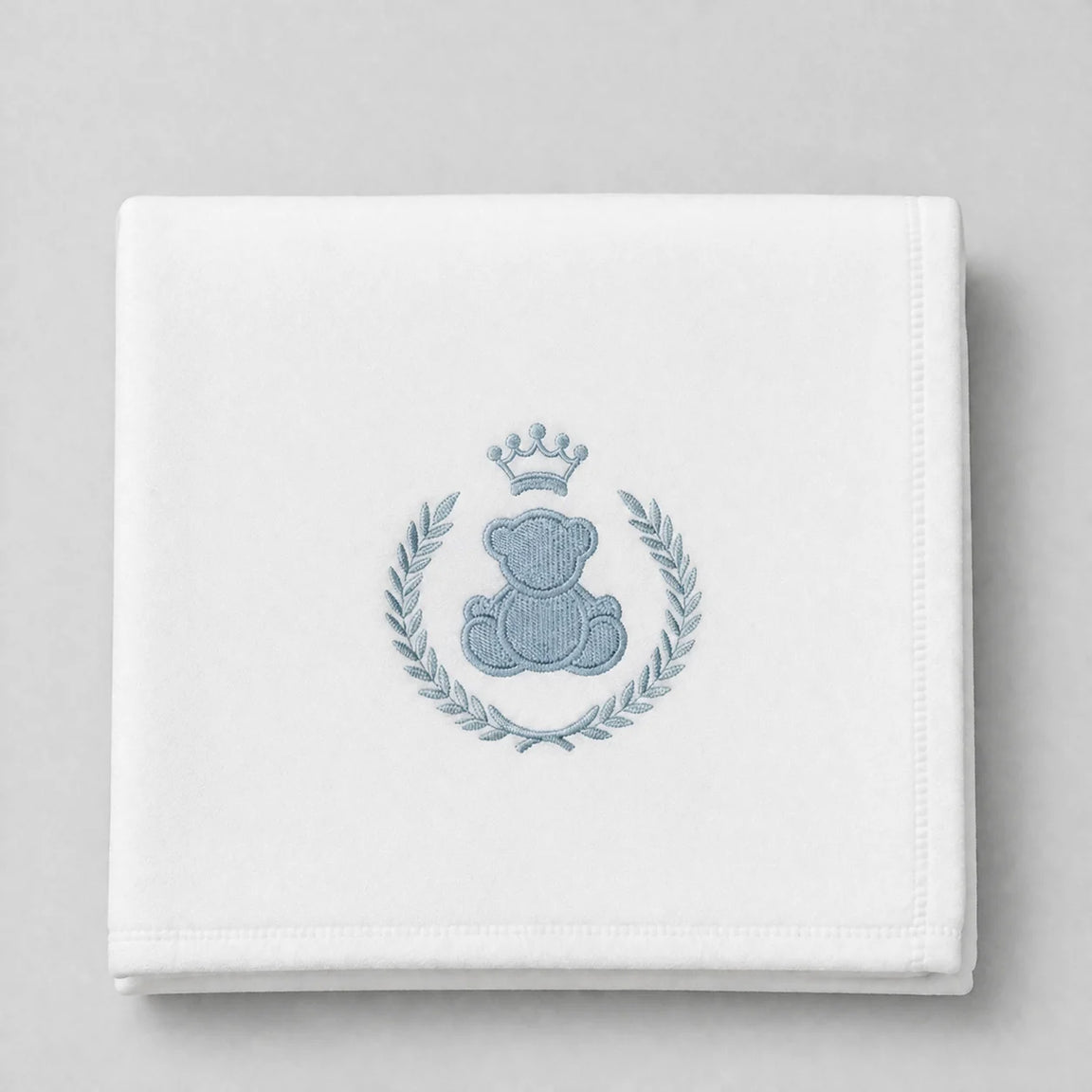 White blanket folded flat with a crowned blue teddy bear and leafy wreath motif