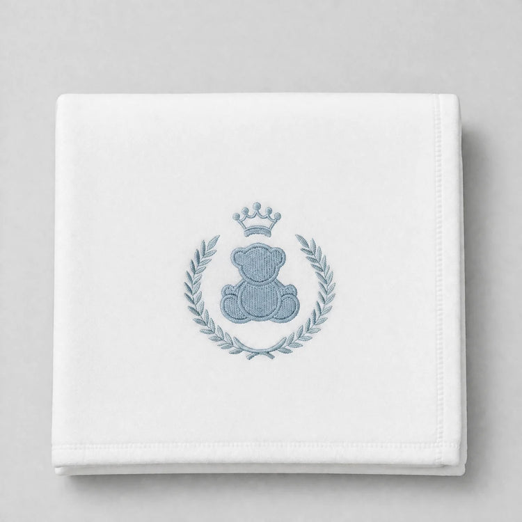 White blanket folded flat with a crowned blue teddy bear and leafy wreath motif