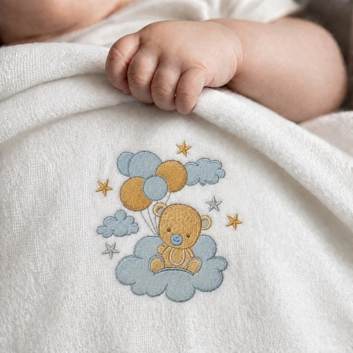 Close-up of an infant's hand on a white blanket with a teddy bear, balloons and clouds
