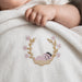 Close-up of an infant's hand on a white blanket with a sleeping baby and floral wreath motif