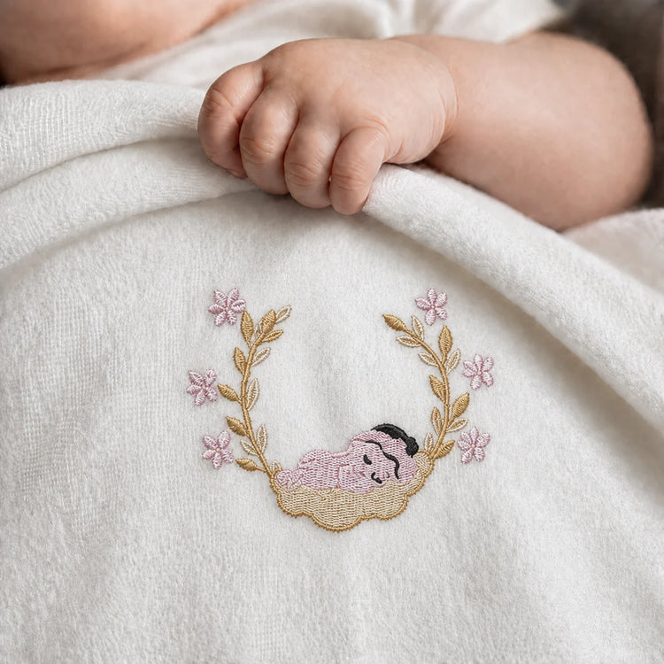 Close-up of an infant's hand on a white blanket with a sleeping baby and floral wreath motif
