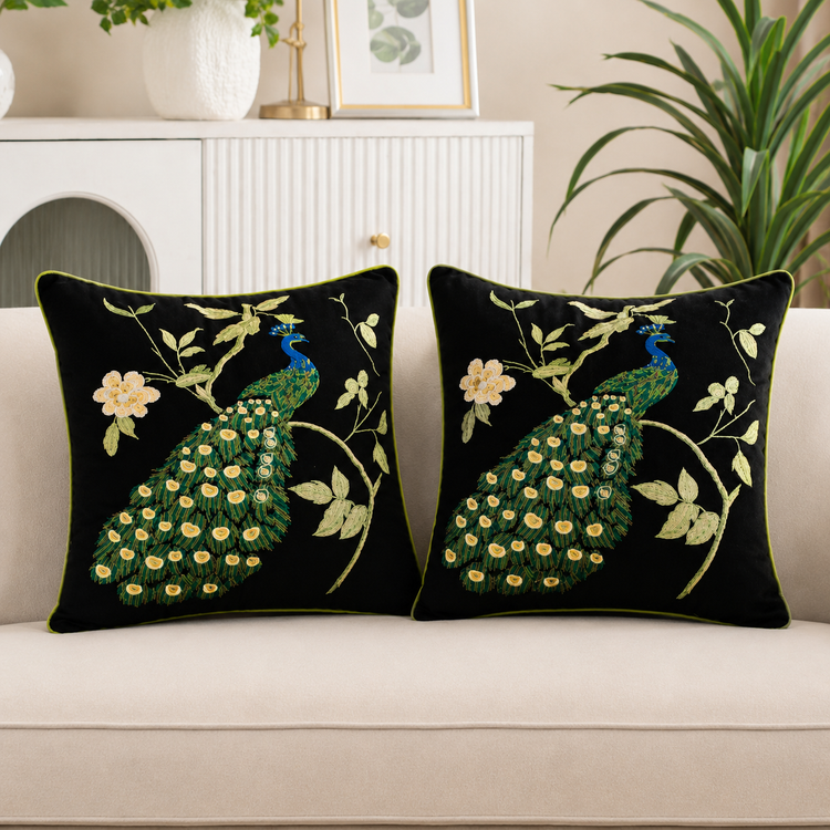 Two black cushion covers with green peacocks and cream flowering branches on a sofa.