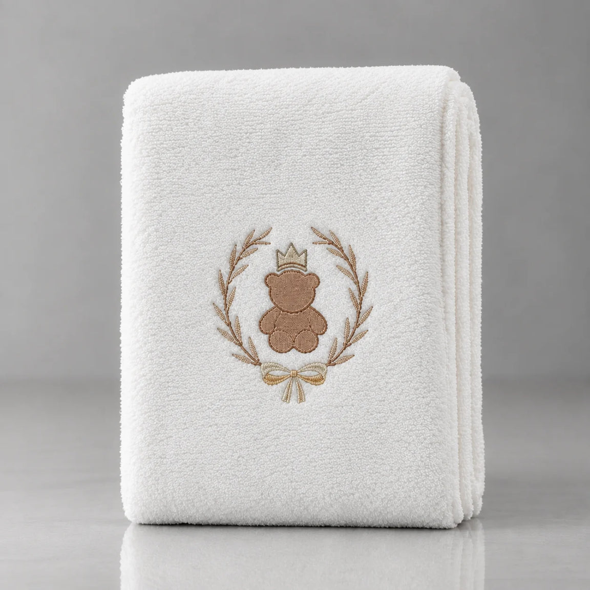 Folded white blanket with a crowned brown teddy bear inside a gold-tone leafy wreath