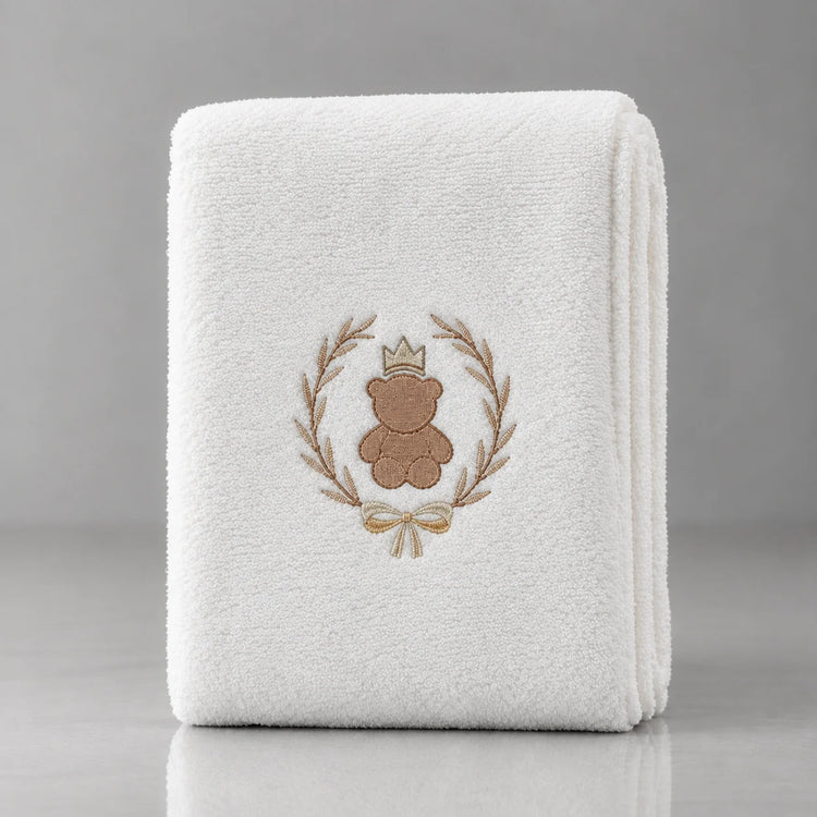 Folded white blanket with a crowned brown teddy bear inside a gold-tone leafy wreath