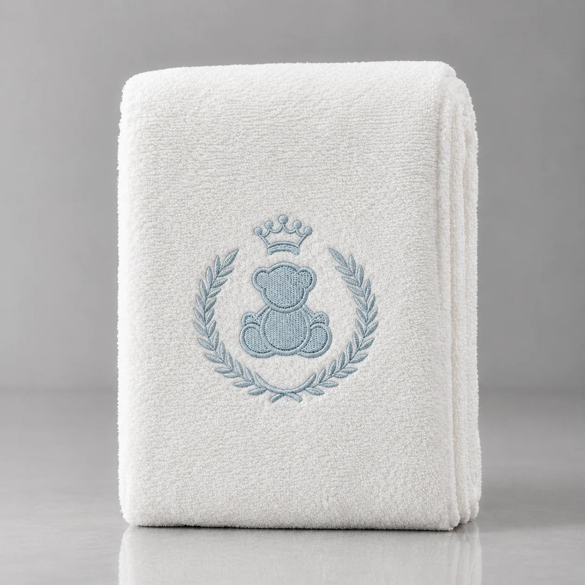 Folded white blanket with a crowned blue teddy bear inside a blue leafy wreath