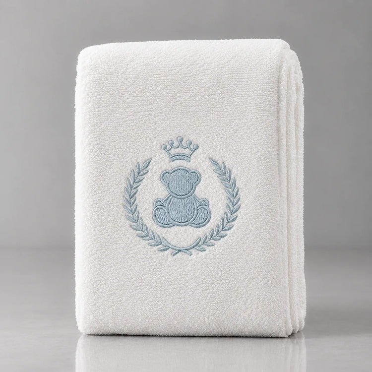 Folded white blanket with a crowned blue teddy bear inside a blue leafy wreath