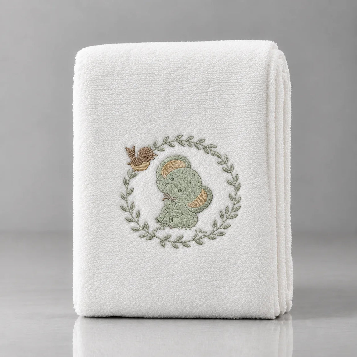Folded white blanket with a pale green elephant inside a leafy wreath