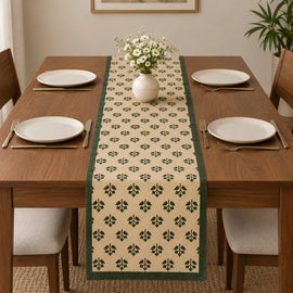 Alt Text: Wooden dining table set with white plates and cutlery, featuring a cream-colored table runner covered in a small, repeating green botanical clover pattern and solid dark green side borders.   