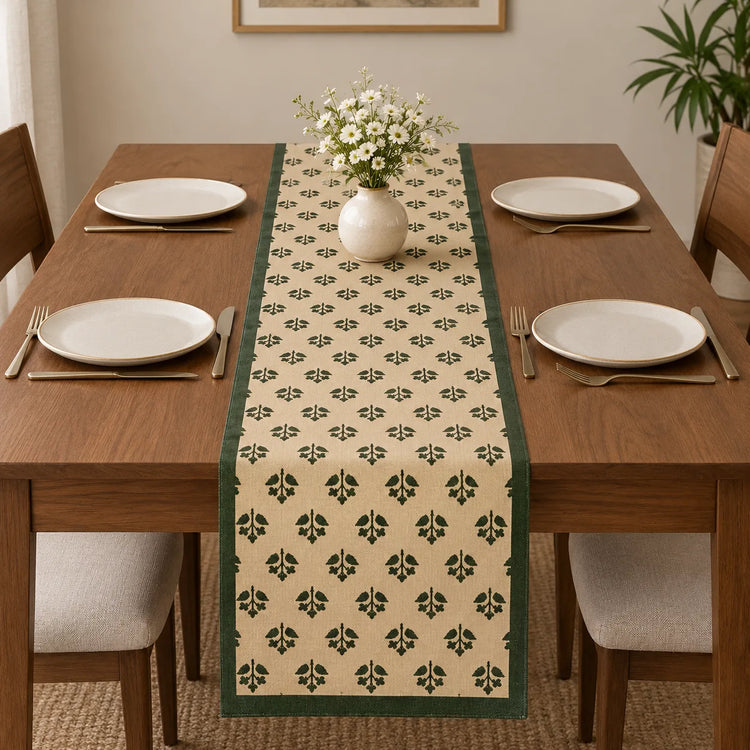 Alt Text: Wooden dining table set with white plates and cutlery, featuring a cream-colored table runner covered in a small, repeating green botanical clover pattern and solid dark green side borders.   