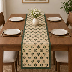 Alt Text: Wooden dining table set with white plates and cutlery, featuring a cream-colored table runner covered in a small, repeating green botanical clover pattern and solid dark green side borders.   