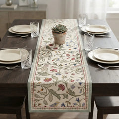A rectangular table runner with an intricate floral and bird pattern in muted red, blue, green, and beige tones, styled on a dark wood dining table with place settings and a small succulent.