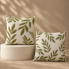 Two square throw pillows with an olive green leaf pattern on a cream linen background, styled on a beige pedestal and against a beige wall with soft leaf shadows.
