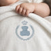 Close-up of an infant's hand resting on a white blanket with a blue teddy bear and wreath motif