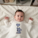 Baby lying beneath an ivory blanket with a blue elephant holding a heart-shaped balloon.