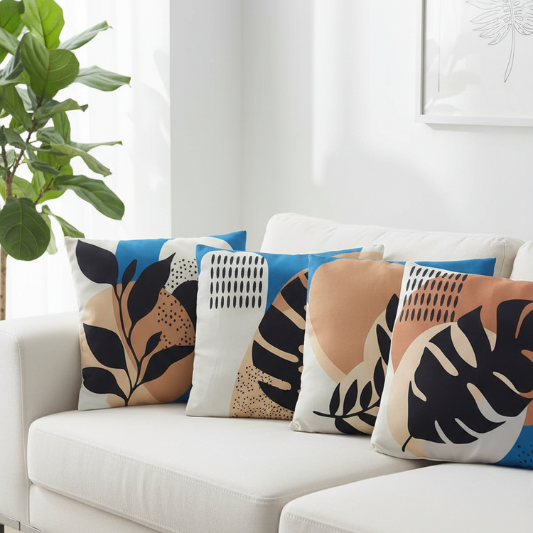 Four cushion covers with black tropical leaves over blue and peach abstract panels on a sofa.