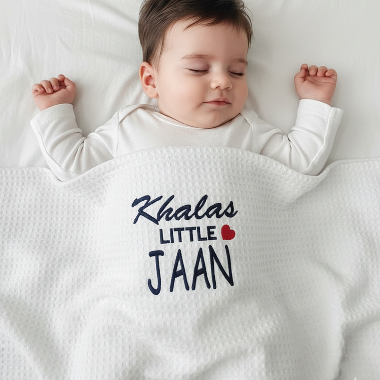 Infant under a white waffle-textured blanket with Khalas Little Jaan lettering and a red heart