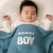 Infant under a blue waffle-textured blanket with white mamas BOY lettering