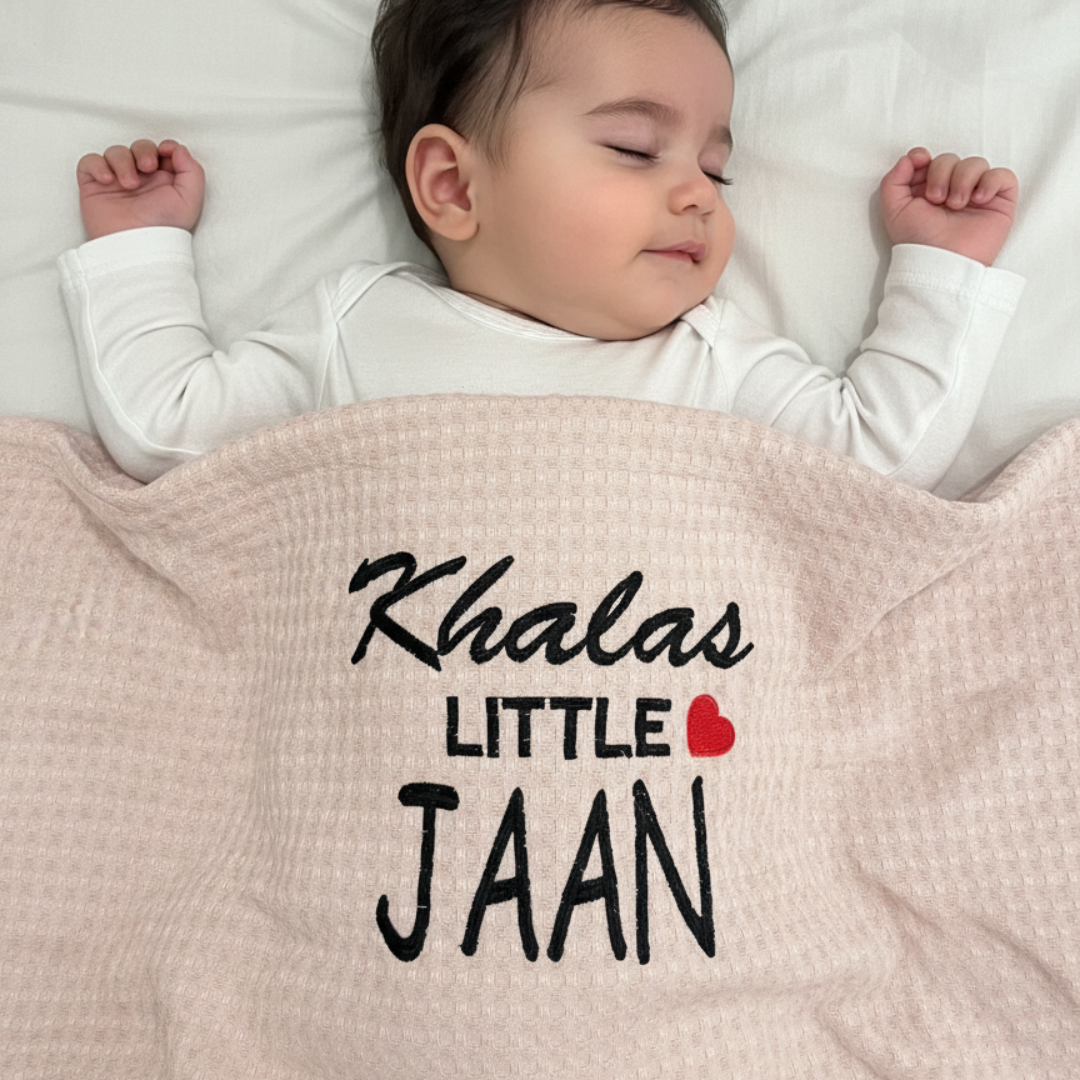 Infant under a beige waffle-textured blanket with Khalas Little Jaan lettering and a red heart