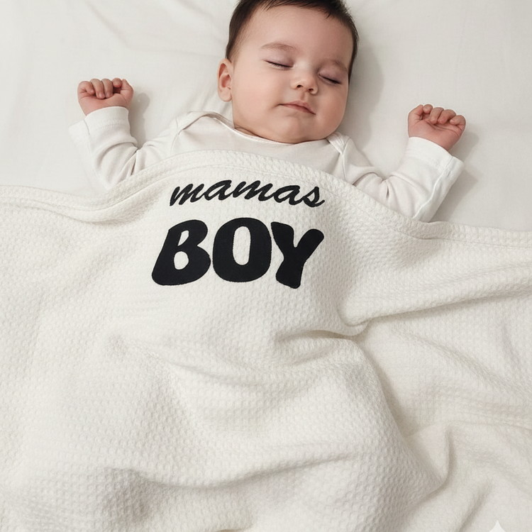 Infant under a white waffle-textured blanket with black mamas BOY lettering