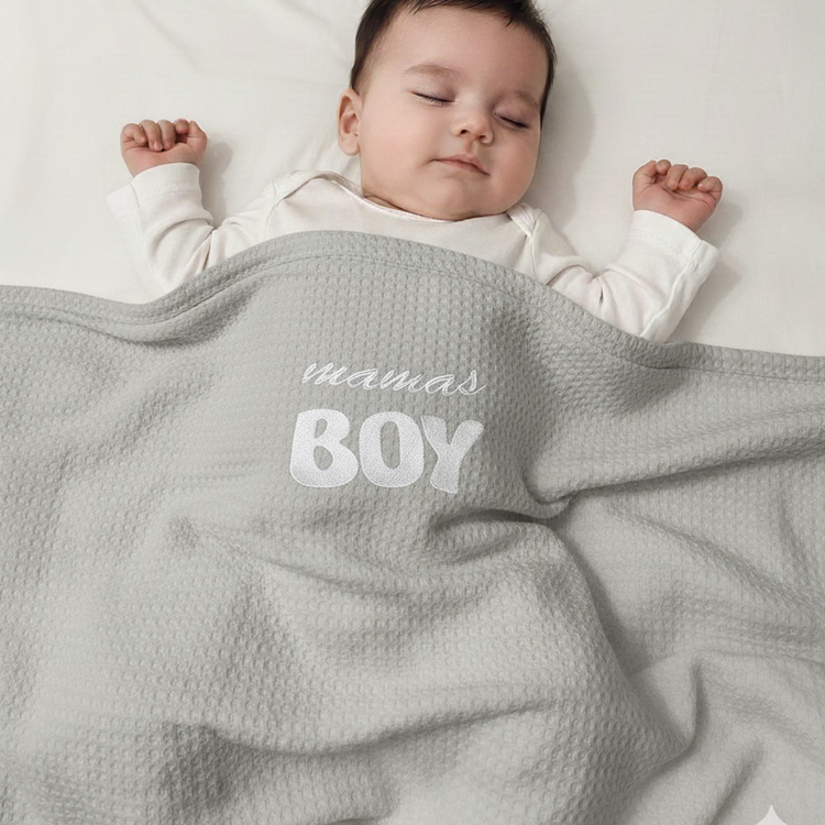 Infant under a gray waffle-textured blanket with white mamas BOY lettering
